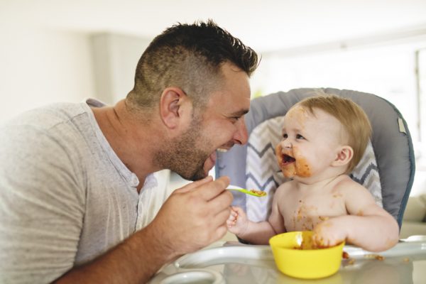 A Little baby eating her dinner and making a mess with dad on the side