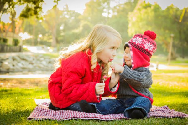 Little Girl with Baby Brother Wearing Coats and Hats Outdoors sharing candy