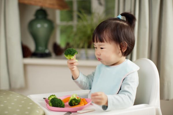 Toddler girl eating healthy vegetable sitting on high chair beside a dinner table