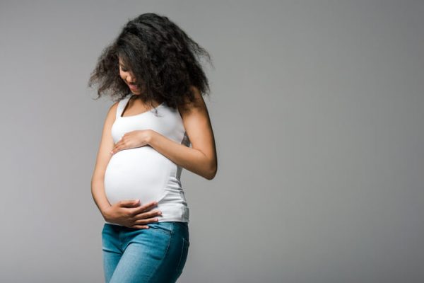 Happy pregnant african american girl touching belly while standing on grey