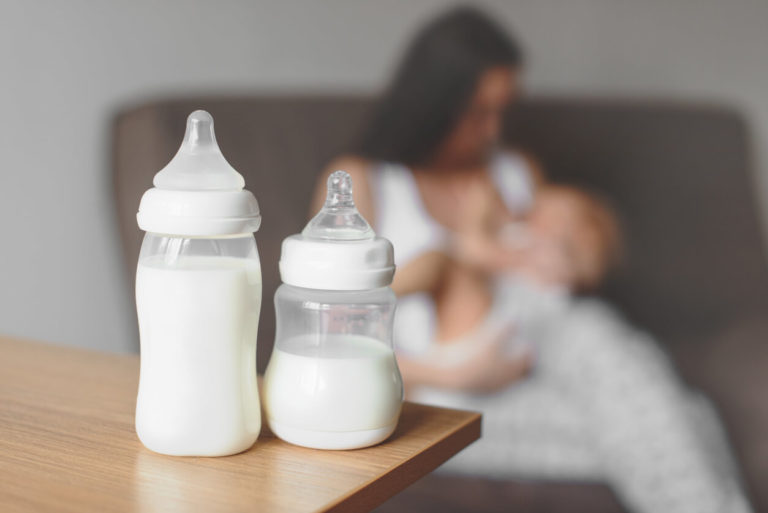 Bottles with breast milk on the background of mother holding in her hands and breastfeeding baby