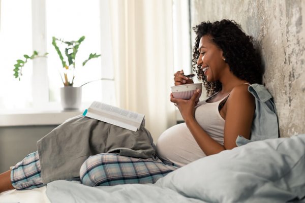 Smiling pregnant woman having breakfast in bed while reading a parenting book