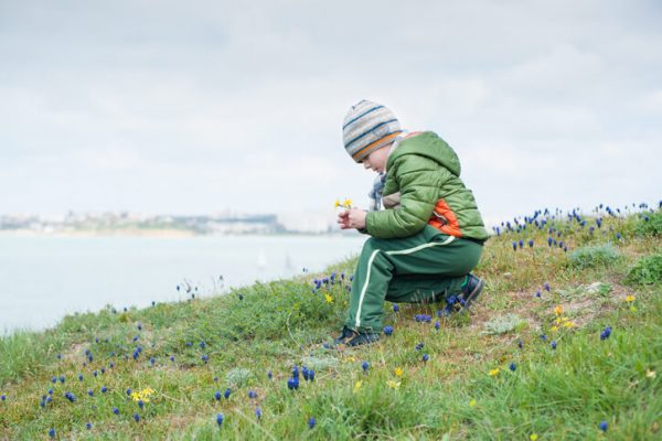 Little child in jacket and hat collecting spring flowers on green slope against backdrop of sea