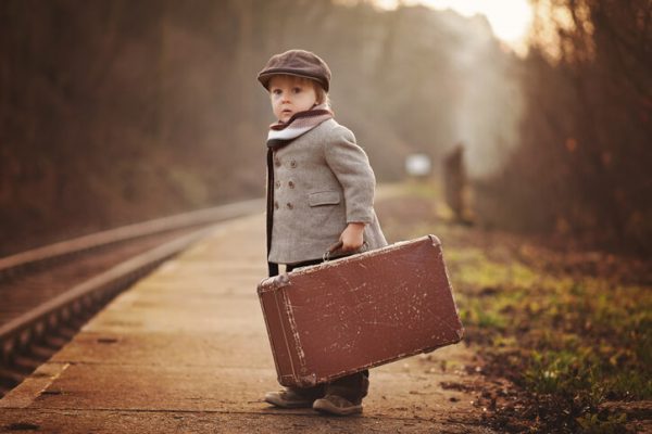 Adorable boy on a railway station, waiting for the train with suitcase and teddy bear